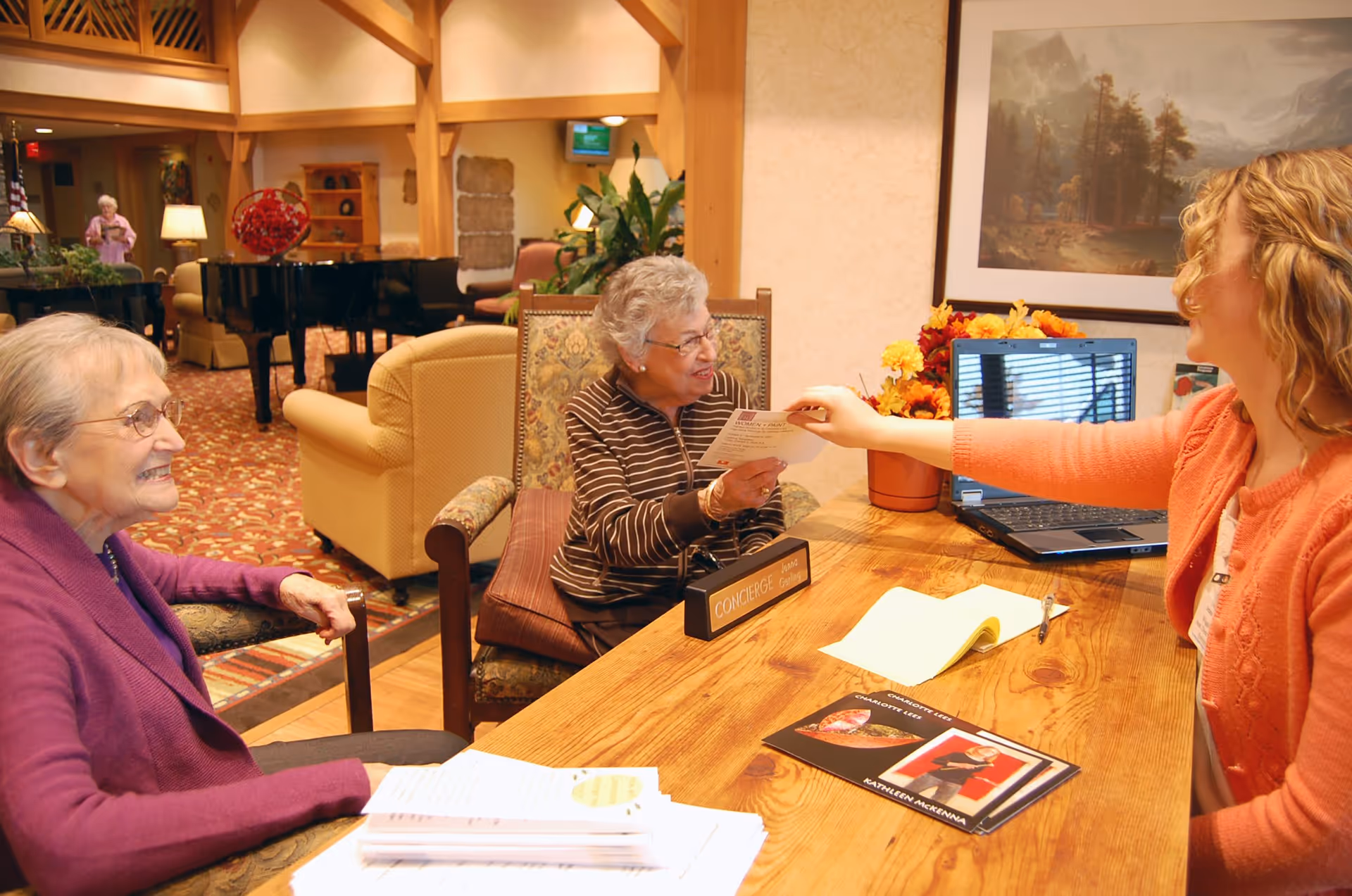 A senior living facility concierge desk where a woman in an orange sweater is handing a paper to an elderly woman sitting behind the desk with a 'CONCIERGE' nameplate. Another elderly woman in a purple sweater is seated nearby, smiling. The background shows a cozy common area with a grand piano, armchairs, plants, and warm lighting.