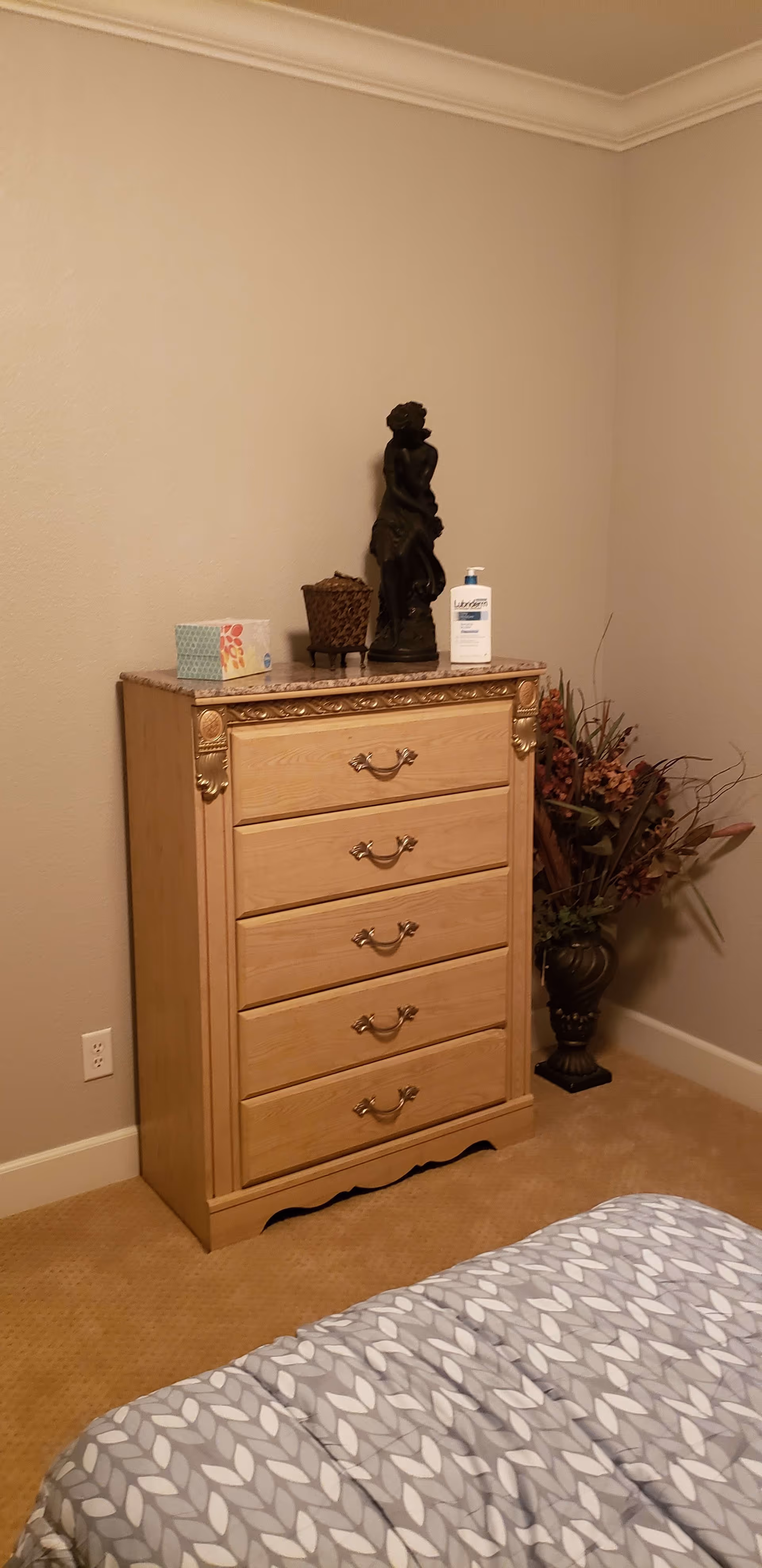 A corner of a bedroom featuring a wooden chest of drawers with ornate handles and decorative trim. On top of the chest are a tissue box, a small basket, a lotion bottle, and a dark statue. To the right of the chest is a large vase with dried flowers. Part of a bed with a patterned gray and white comforter is visible in the foreground. The walls are beige, and the floor is carpeted.