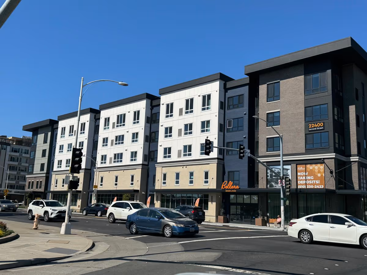 Facade of Bellara Senior Living, a multi-story building with street-level storefronts and cars at an intersection under a clear blue sky.