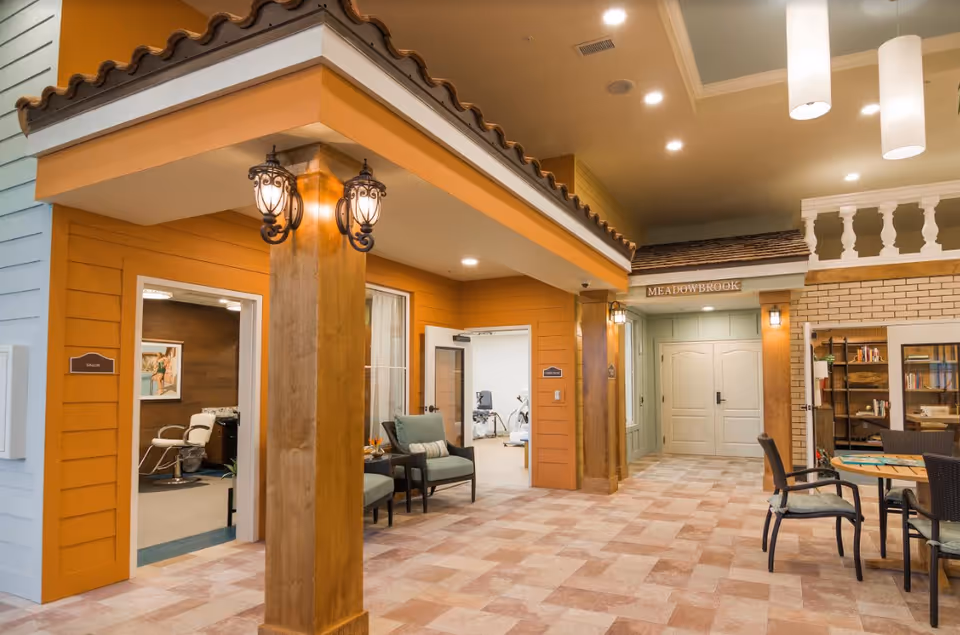 Interior common area of a memory care facility styled like a small village with storefront facades, seating, and a reading table.