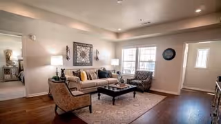 A well-lit living room with a beige sofa, patterned armchair, coffee table, and decorative wall art. The room has hardwood floors, a large window letting in natural light, and a doorway leading to a bedroom.