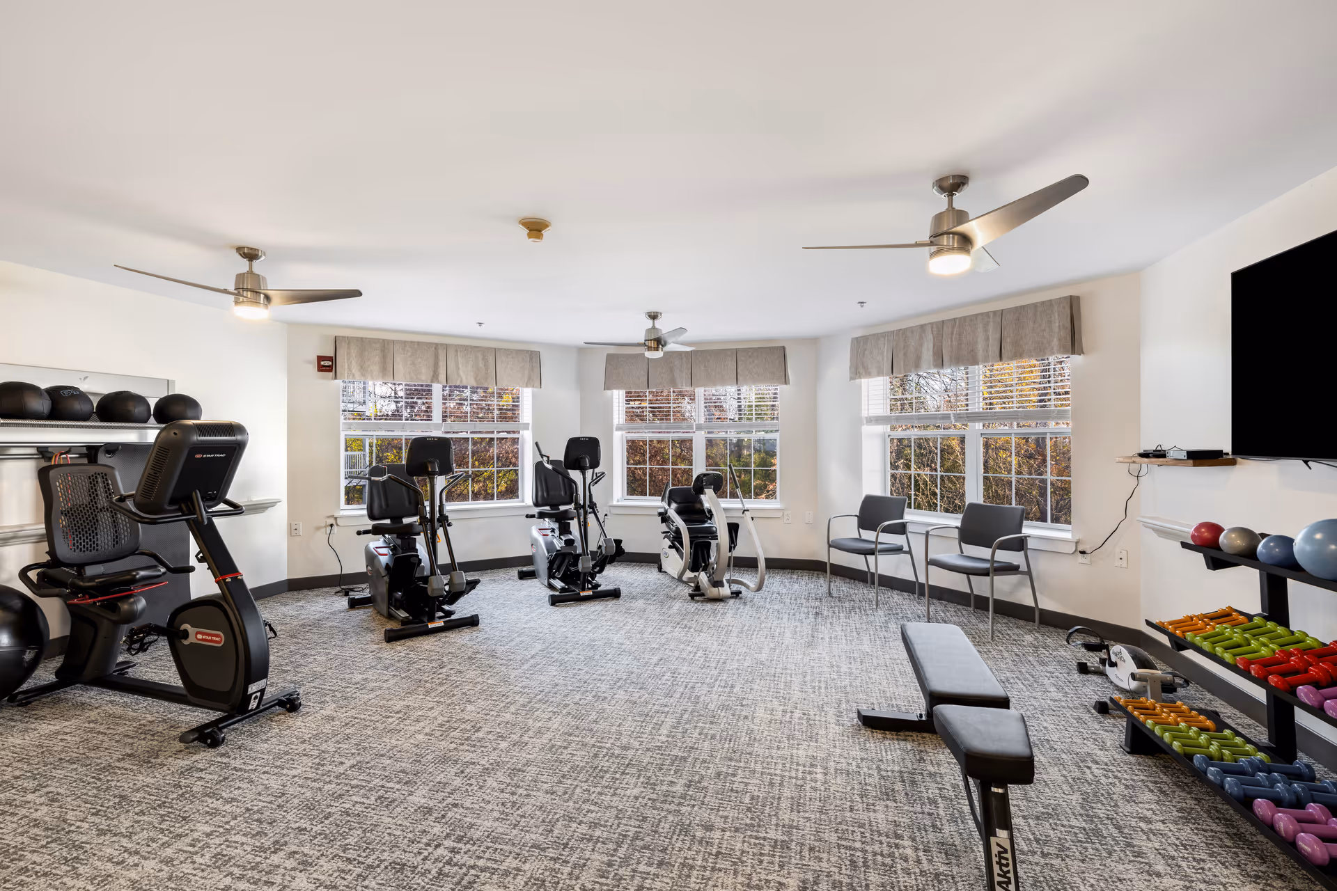 A bright fitness room with large windows letting in natural light. The room contains several exercise machines including stationary bikes and a rowing machine, a bench, and shelves with various colorful dumbbells and exercise balls. There are also three chairs lined up near the windows and ceiling fans overhead.