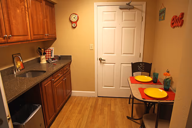 Small kitchen area with wooden cabinets, a granite countertop with a sink, and a small dining table set for two with yellow plates and blue glasses. The room has wooden flooring, a white door, a wall clock, and decorative signs that say 'coffee' and 'eat'.