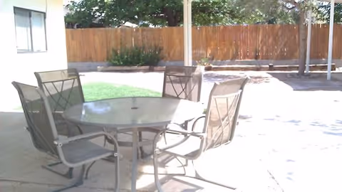 Outdoor patio area with a round glass-top table and four metal mesh chairs. The area is shaded by a roof and surrounded by a wooden fence with some greenery visible in the background.