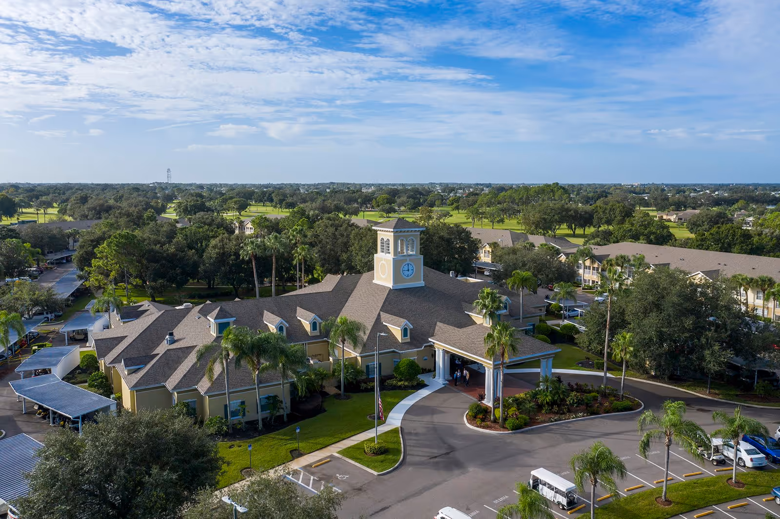 Aerial view of a senior living facility building with a clock tower, surrounded by palm trees and greenery, with parking areas and golf course in the background under a partly cloudy sky.
