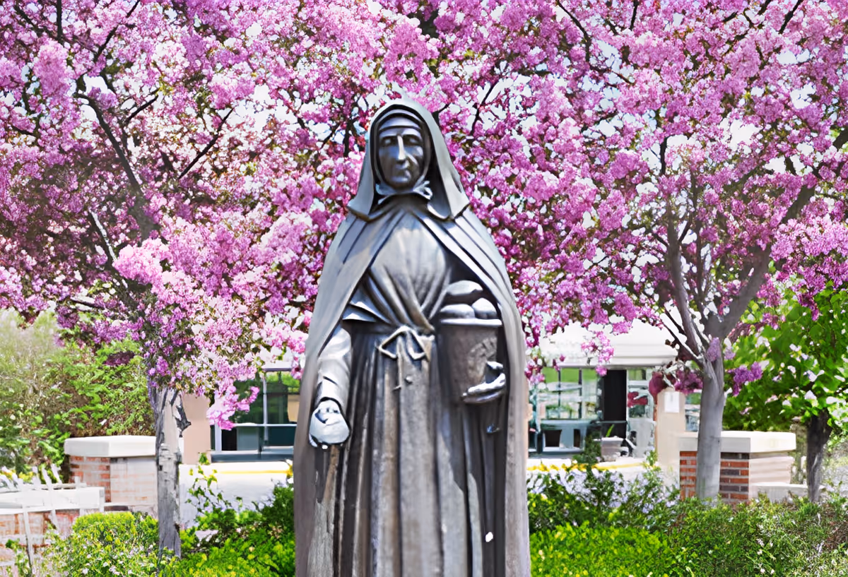 Bronze statue of a nun holding a basket set before pink flowering trees and a building entrance.