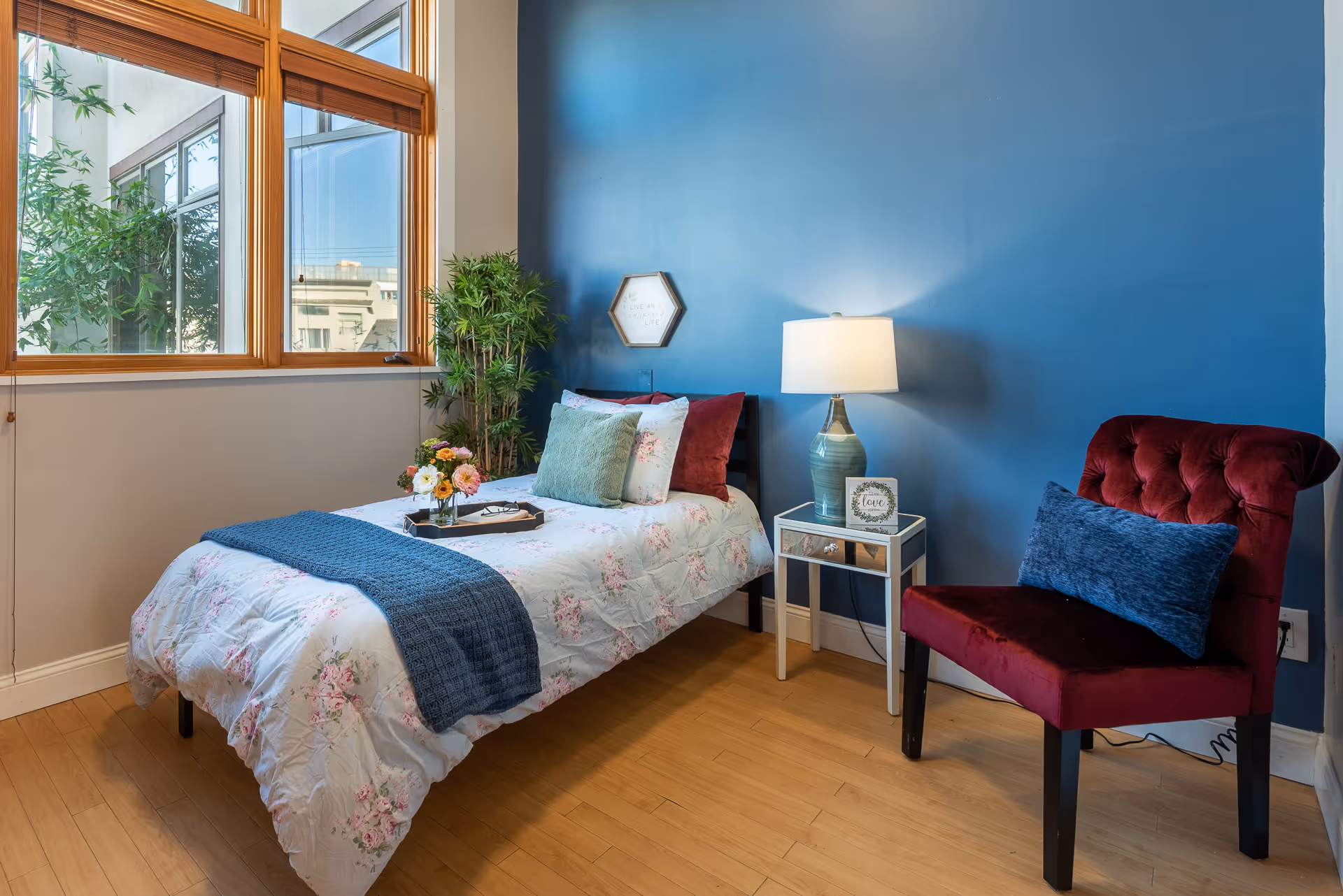A cozy bedroom with a single bed covered in floral bedding and a blue throw blanket. There are multiple pillows on the bed, including green and burgundy ones. Next to the bed is a small mirrored nightstand with a blue lamp and a decorative sign. A burgundy upholstered chair with a blue cushion is placed against a blue accent wall. Large windows let in natural light, and a green potted plant is near the bed.