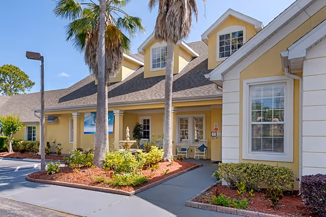 Exterior view of a yellow building with white trim, featuring a covered entrance with two palm trees and a small garden with shrubs and a fountain in front. There is a parking area and a street lamp visible on the left side under a clear blue sky.