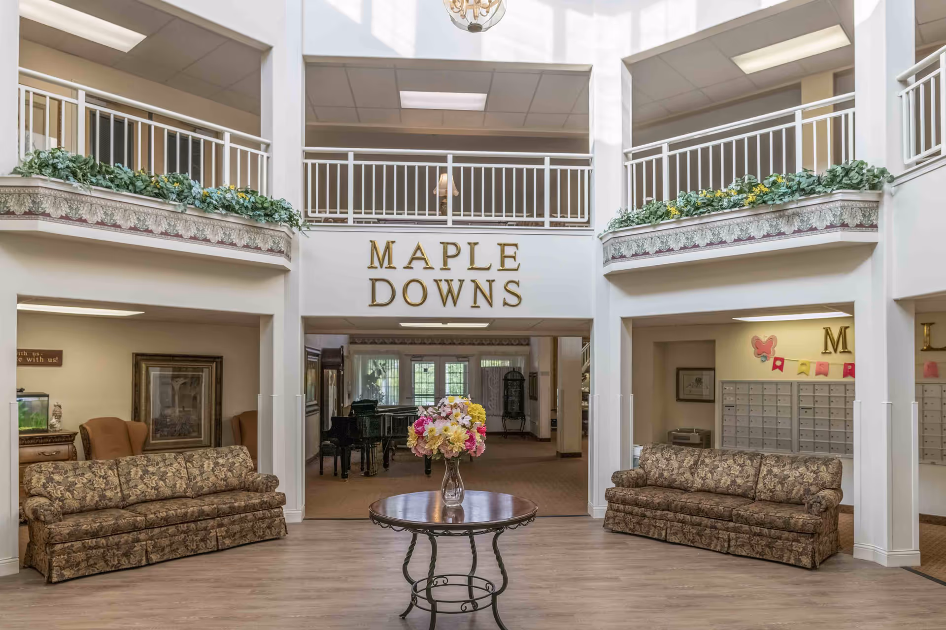 Interior view of the Holiday Maple Downs facility showing a spacious lobby area with two patterned sofas on either side, a round table with a vase of flowers in the center, and a balcony with white railings above. The words 'MAPLE DOWNS' are displayed prominently on the wall above the entrance to another room with a piano visible inside.