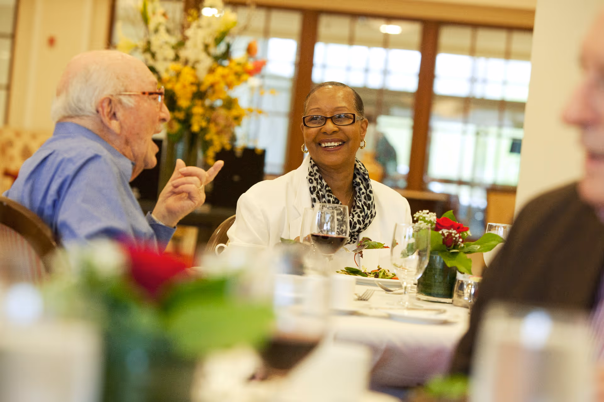 Two elderly people sitting at a dining table in a well-lit room, engaged in conversation and smiling. The table is set with glasses, plates, and a small floral centerpiece. There are large windows and a vase with yellow flowers in the background.