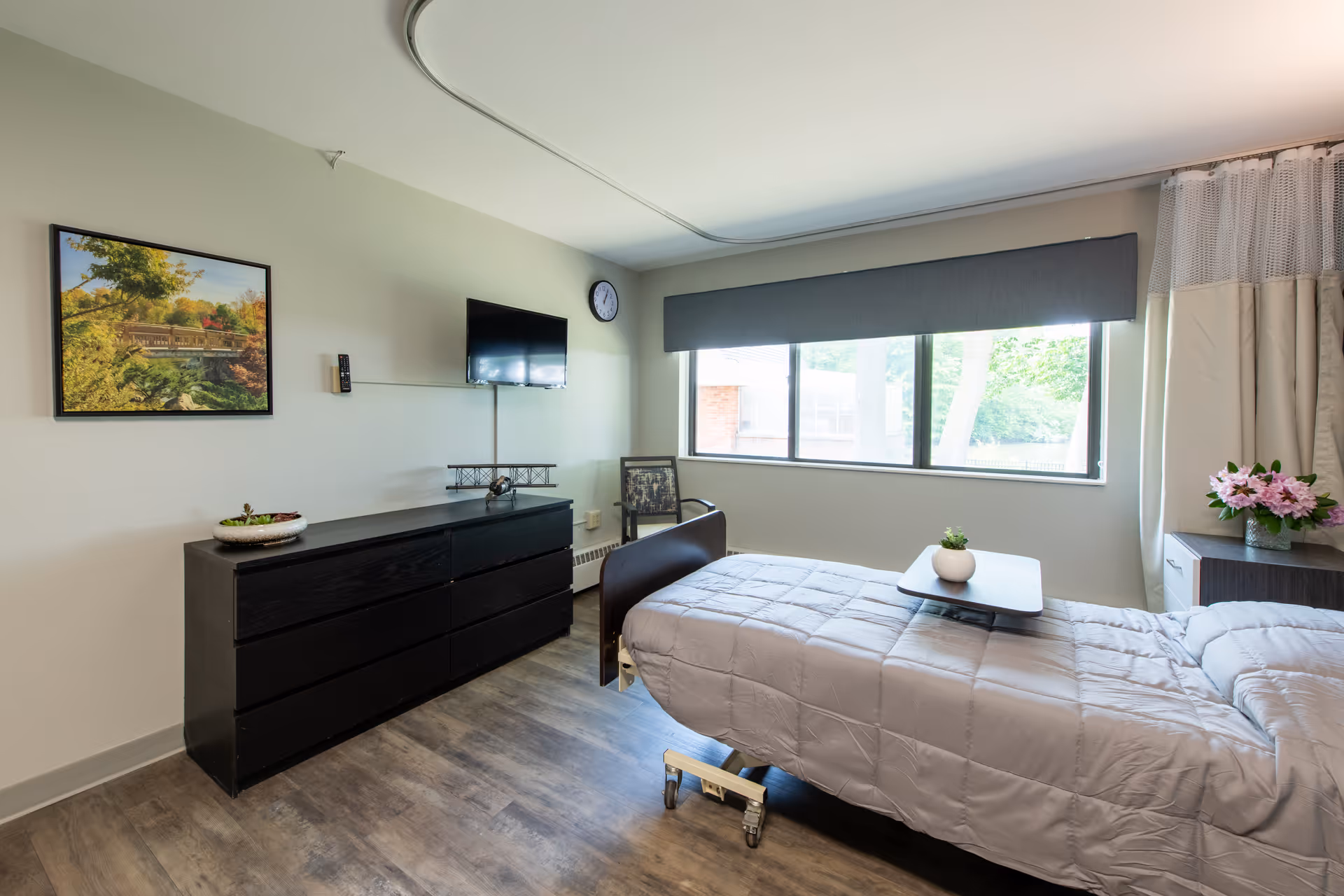A bright senior living bedroom with a hospital-style bed, low dresser, wall-mounted TV and a large window.