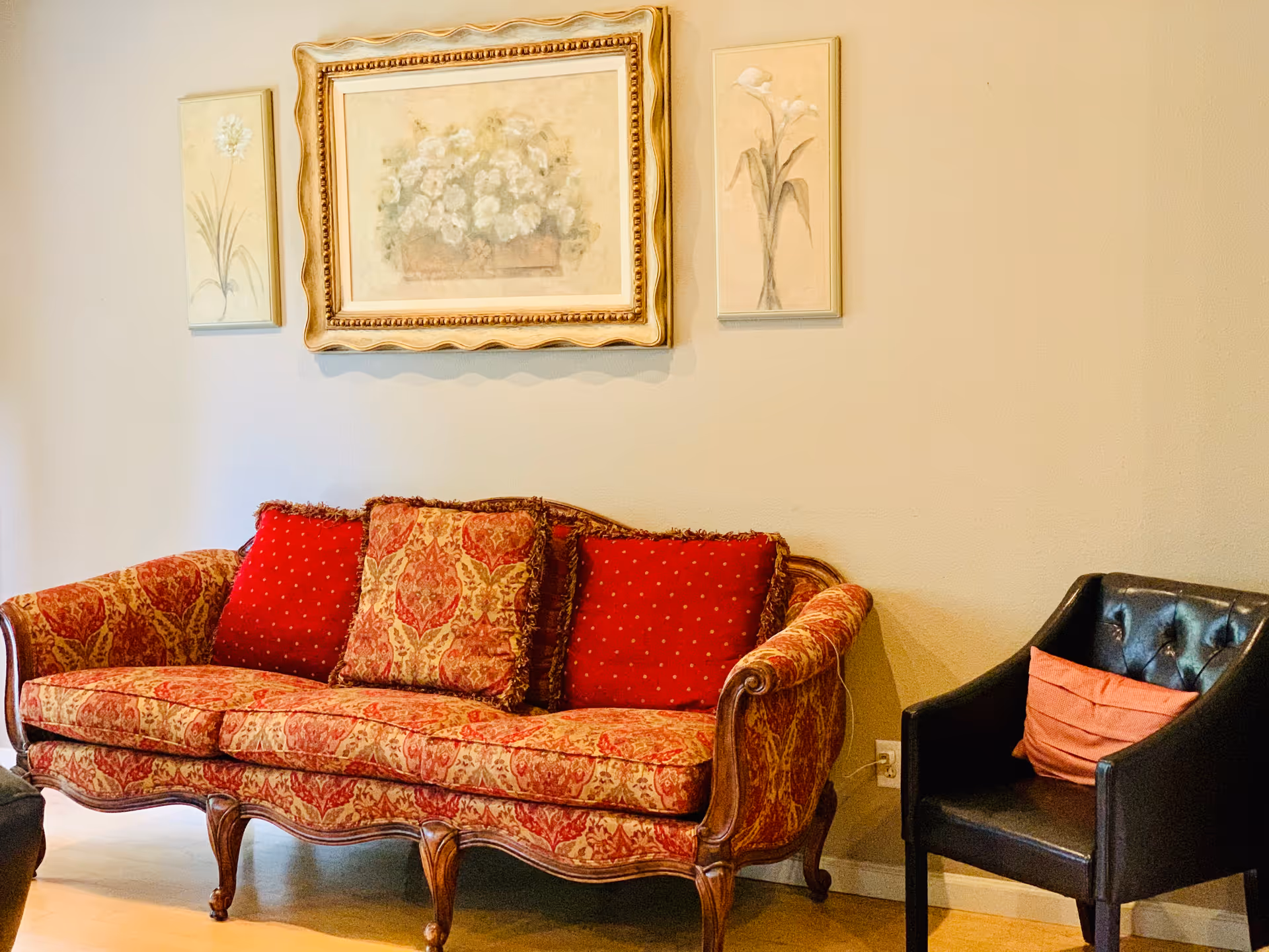 A cozy living room area featuring a vintage-style patterned sofa with red and gold cushions, a black leather armchair with an orange pillow, and three floral paintings hanging on a beige wall above the sofa.