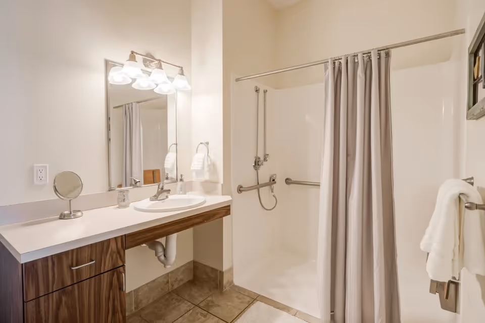 A clean bathroom featuring a vanity with a sink, a large mirror with four light fixtures above it, a small round makeup mirror, and a walk-in shower with grab bars and a gray shower curtain. White towels hang on a towel rack next to the shower.