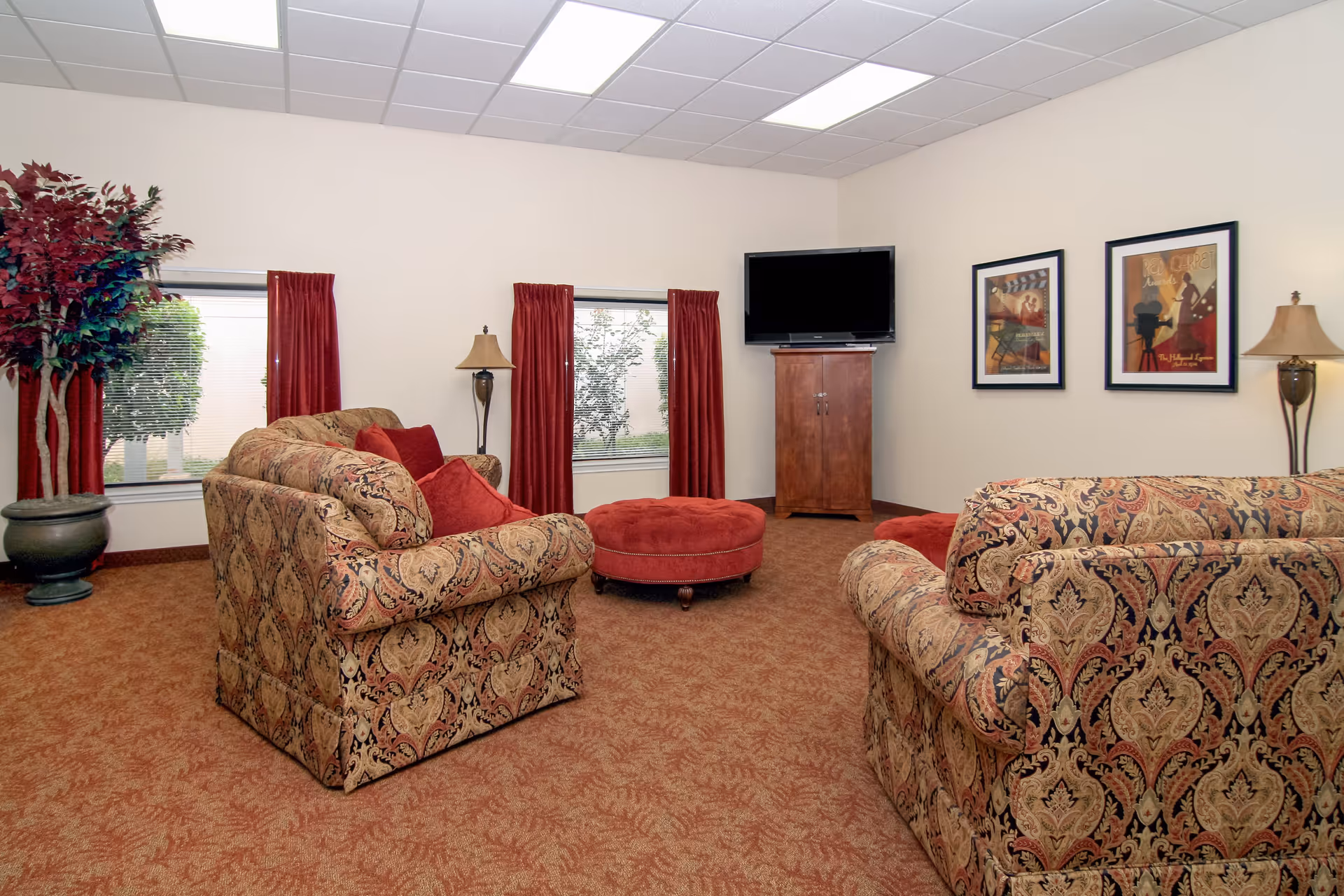 A cozy living room with patterned upholstered sofas, a round red ottoman, two floor lamps, two windows with red curtains, a flat-screen TV on a wooden cabinet, and two framed pictures on the wall.