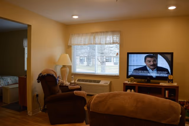 Cozy living room with two recliner chairs, a lamp, a window with blinds above a wall AC/heat unit, and a TV on a stand.