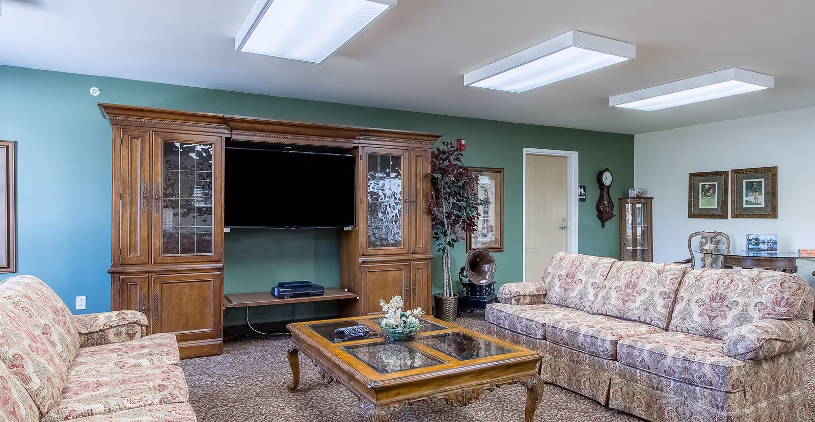Common living room with floral-patterned sofas, a wooden entertainment center with a TV, and a glass-top coffee table.