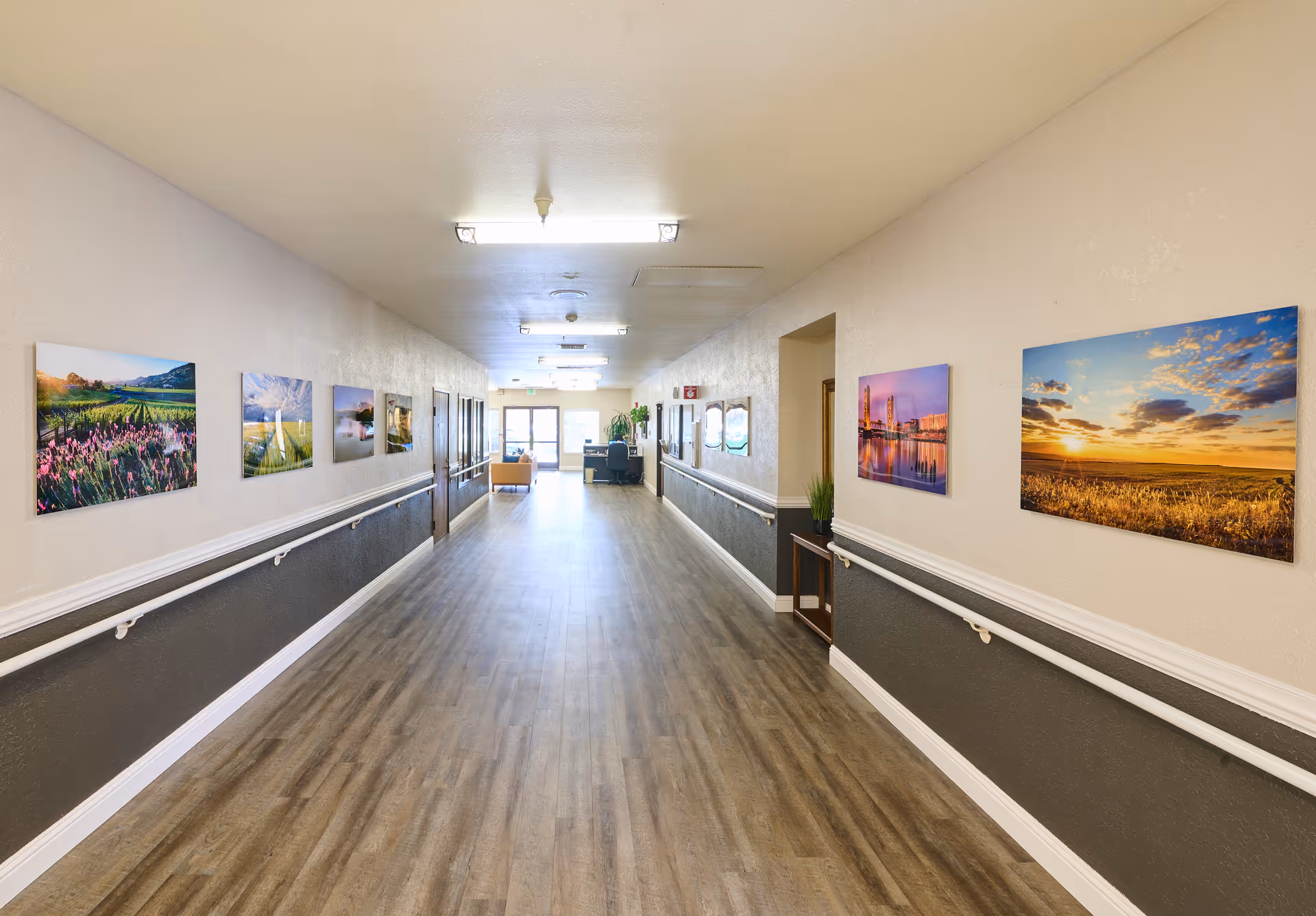 Long, well-lit nursing home hallway with handrails, wood-look flooring and landscape pictures on the walls leading to a seating area and exit.