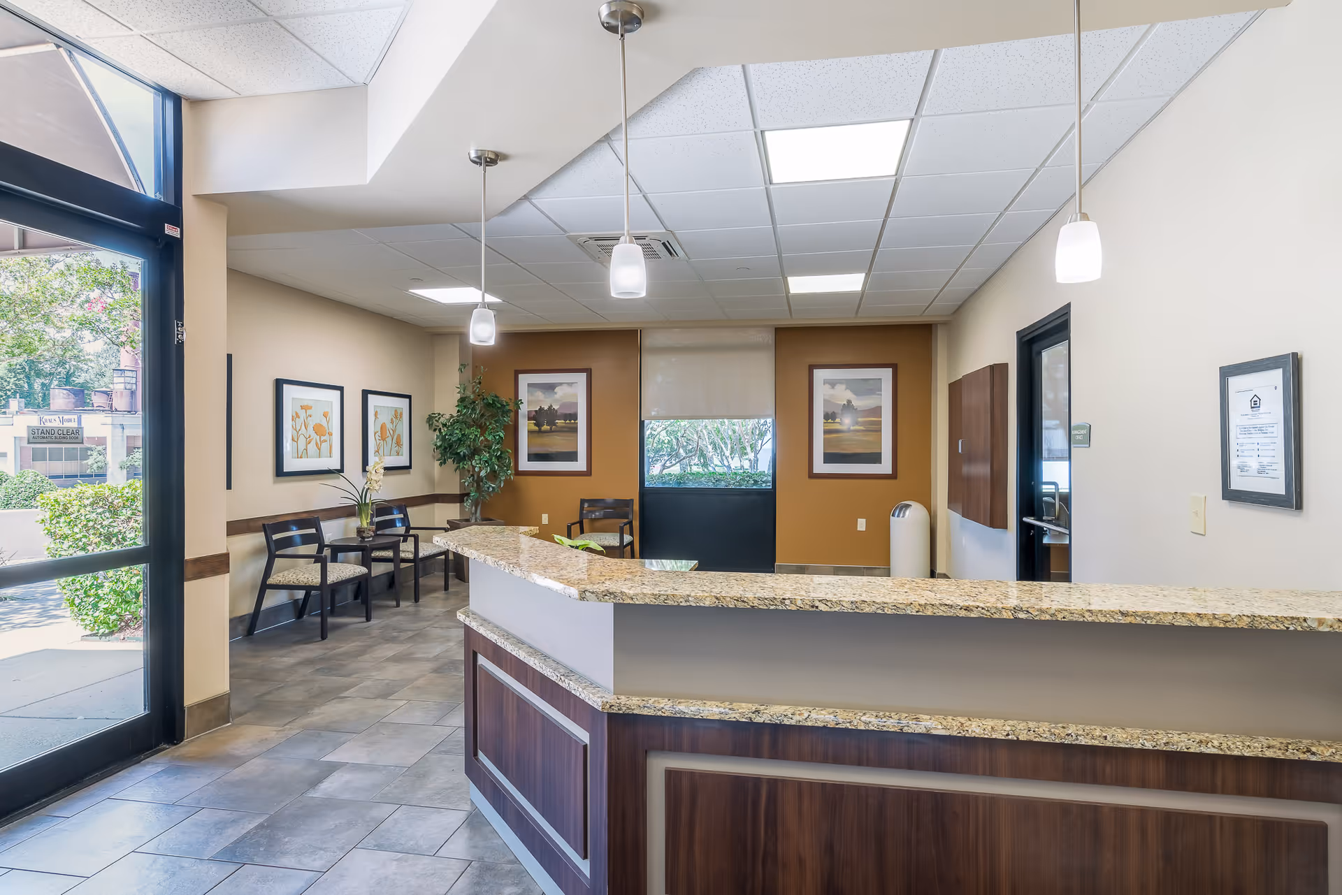 Reception area of City View Towers Senior Apartments featuring a granite countertop desk with pendant lights above, a seating area with chairs and framed artwork on the walls, a large window with a view of greenery outside, and a glass door entrance.