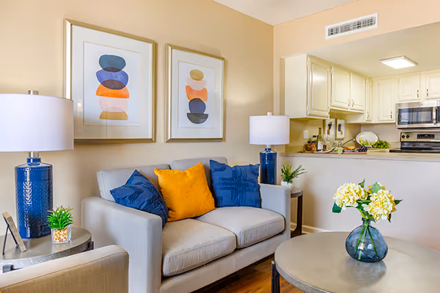 Bright living room with a gray sofa, blue and yellow throw pillows, two table lamps, abstract wall art, a round coffee table with flowers, and a view into a kitchen with a pass-through counter.