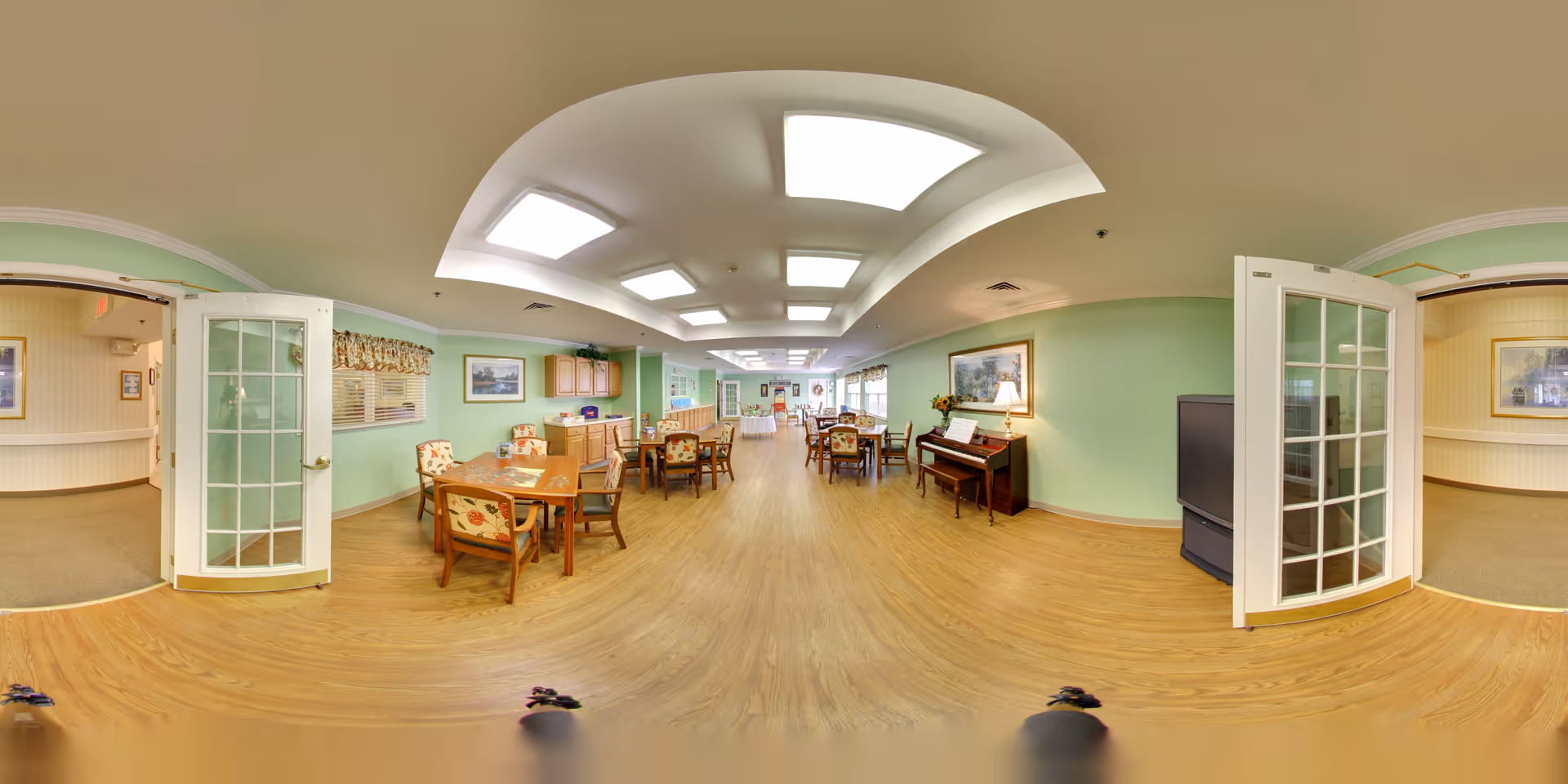 A spacious and well-lit common area in a senior living facility with light green walls and wooden flooring. The room features several wooden tables and chairs with floral upholstery, a piano with a lamp on top, framed artwork on the walls, and large ceiling lights. There are glass-paneled double doors leading to adjacent rooms with carpeted floors.