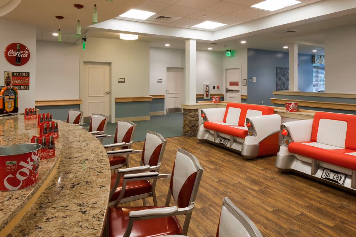 Interior view of a retro-themed lounge area with a curved granite countertop bar, red and white chairs, and vintage Coca-Cola decor. There are two red and white car-style sofas with license plates, wooden flooring, and soft lighting in the ceiling.