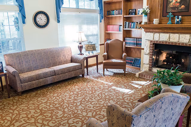 A cozy living room with patterned carpet, a floral sofa, two armchairs, a wooden side table with a lamp, a built-in bookshelf filled with books, and a stone fireplace with a wooden mantle decorated with vases and a framed painting.