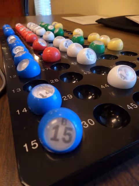 Close-up view of a bingo number holder with colorful numbered balls arranged in rows on a wooden table, with papers and a black folder in the background.