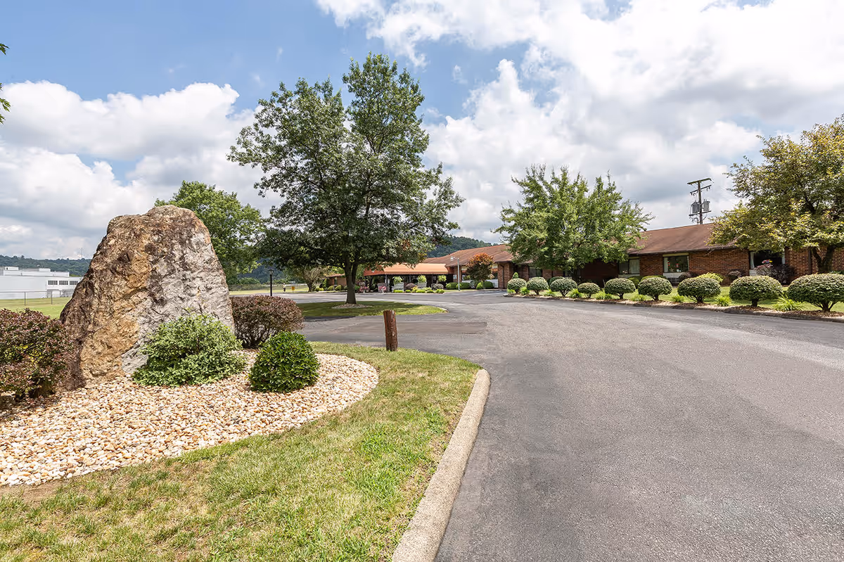 Driveway entrance and landscaped front lawn with shrubs, a large decorative rock, and a single-story brick building under a partly cloudy sky.