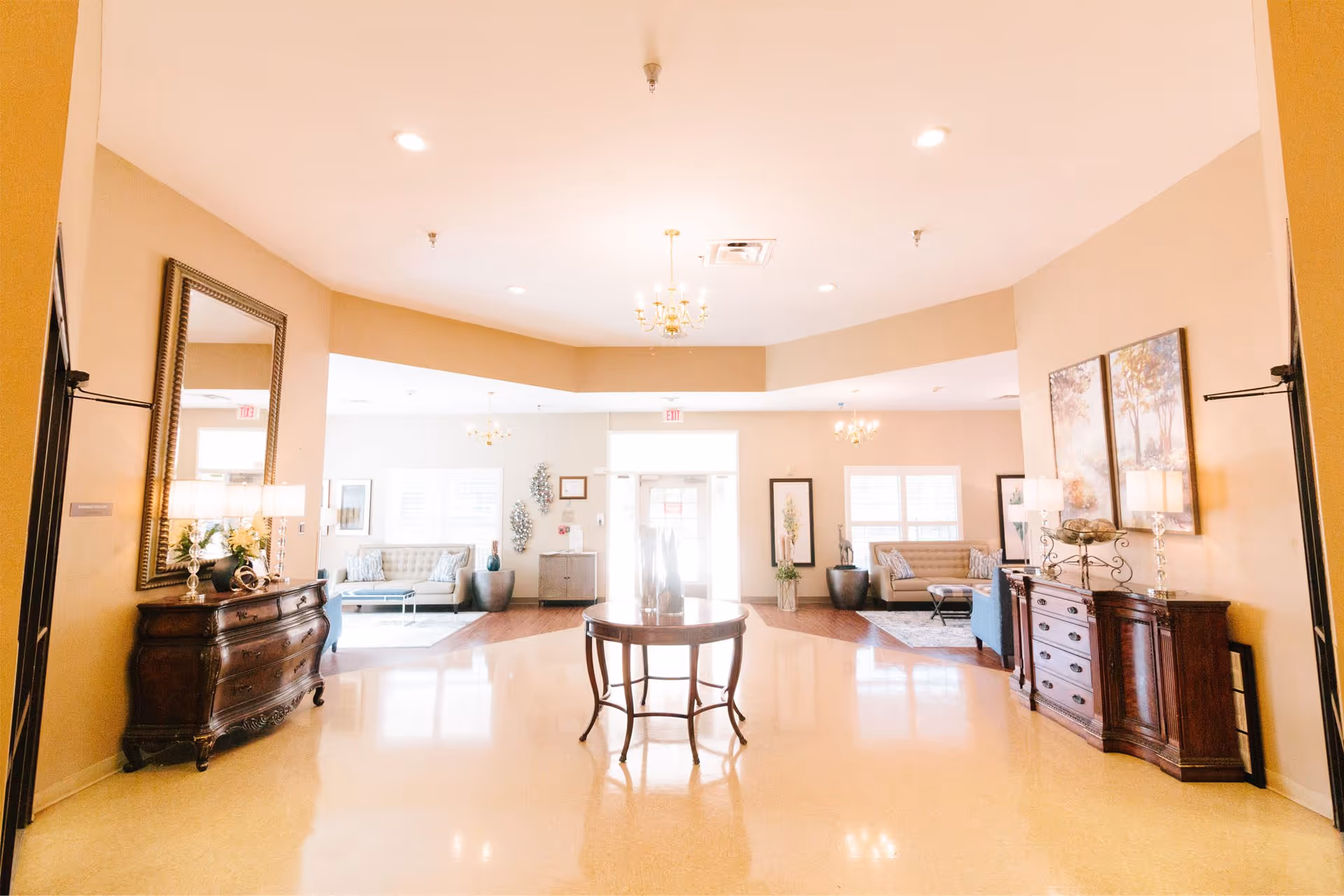 Bright senior living facility lobby with seating areas, decorative dressers, a central round table, and chandeliers.