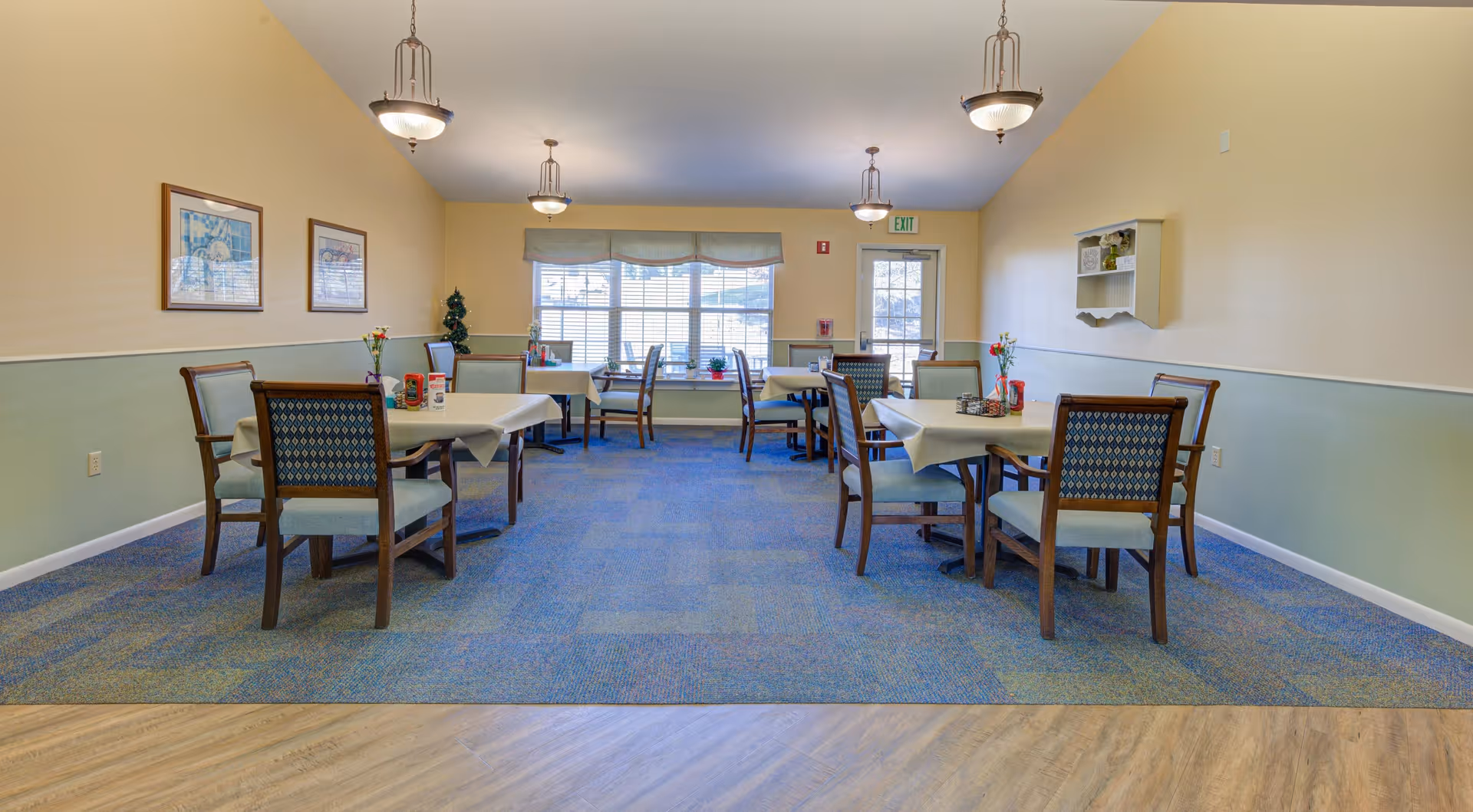 A bright dining room in a senior living community with several tables covered with white tablecloths, each surrounded by wooden chairs with cushioned seats and backs. The room has blue carpet flooring, light yellow and green walls, three hanging light fixtures, framed pictures on the left wall, and a window with a valance at the far end letting in natural light. There is a door next to the window and a small wall-mounted shelf on the right wall.
