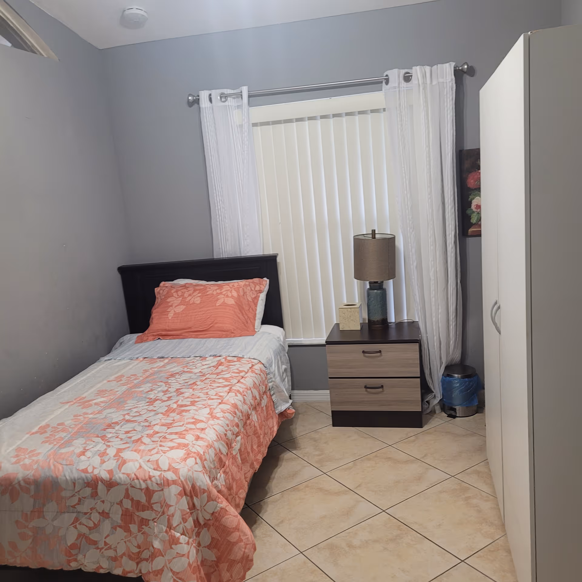 Small single bedroom with a twin bed, floral bedspread, nightstand with a lamp, wardrobe and a window with vertical blinds.