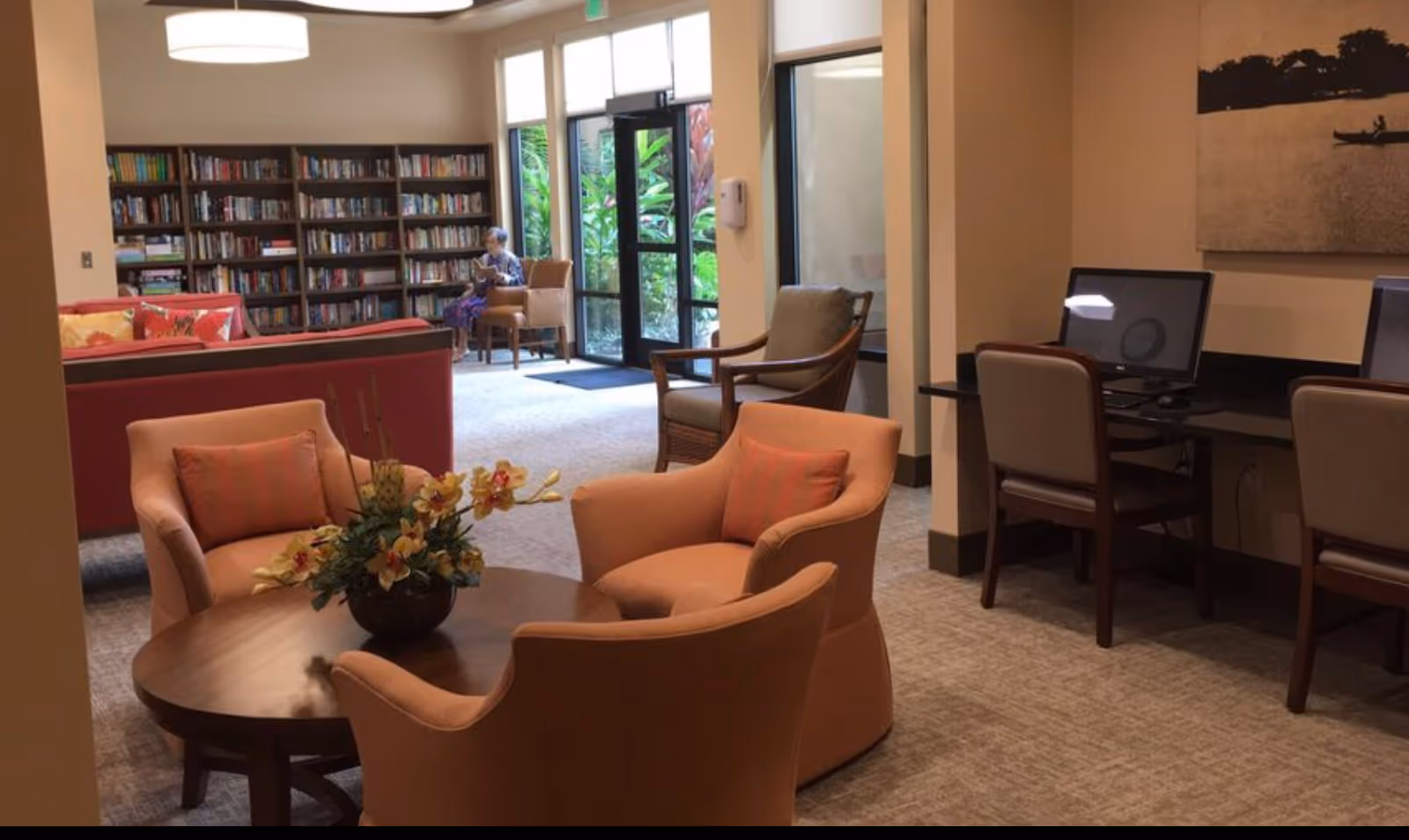 Seating area in a senior living lounge with armchairs around a coffee table, bookshelves in the background, and computer stations to the right.
