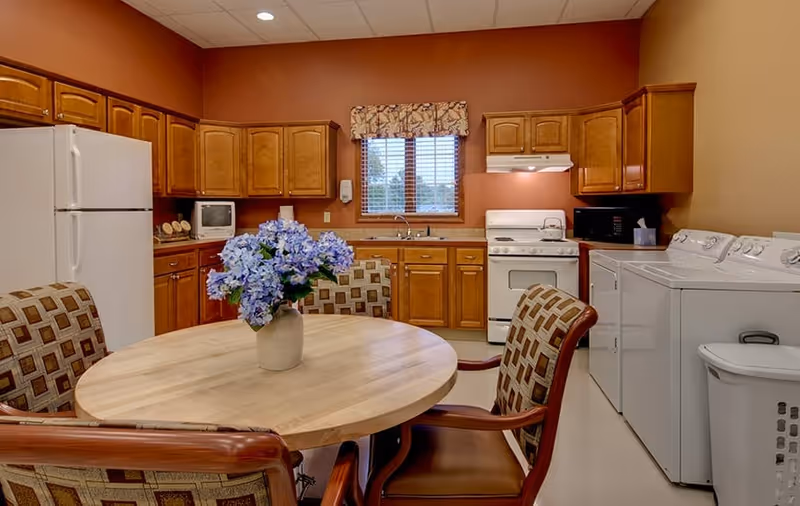 A kitchen and laundry room combination in a senior living facility with wooden cabinets, a white refrigerator, stove, microwave, and a washer and dryer. A round wooden table with four cushioned chairs is in the foreground, with a vase of blue flowers on the table. A window with blinds and a floral valance is above the sink.