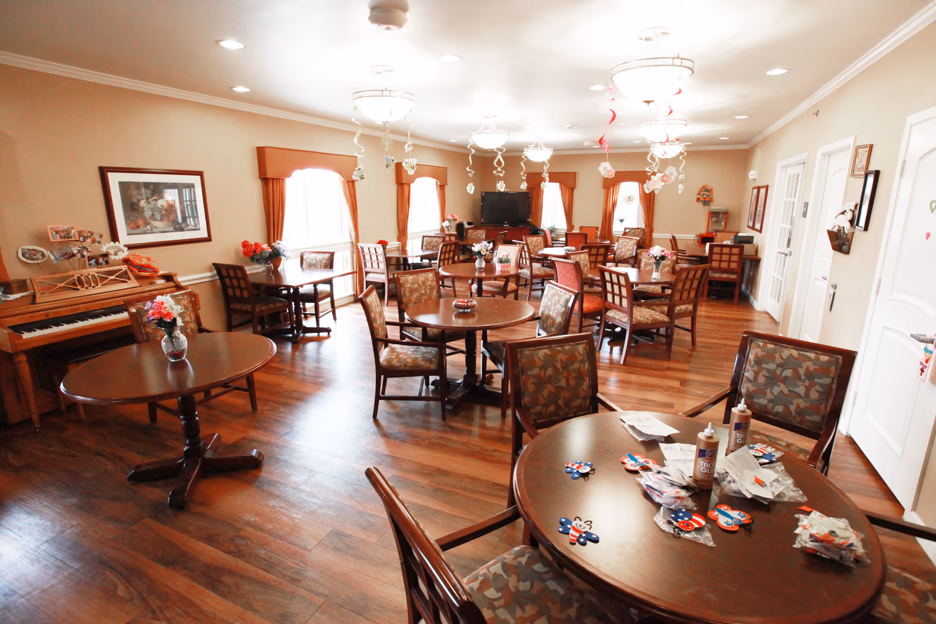A bright and spacious common room with multiple round wooden tables and cushioned chairs arranged neatly. The room features wood flooring, several windows with orange curtains, and a piano against the left wall. Decorative hanging spirals with flowers are suspended from the ceiling. Various craft supplies and decorations are placed on the tables.