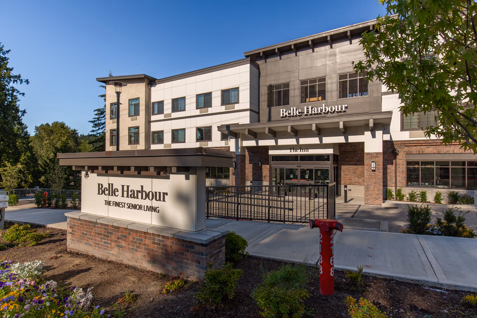 Exterior view of Belle Harbour senior living facility building with a large sign in front that reads 'Belle Harbour THE FINEST SENIOR LIVING'. The building is modern with multiple windows and a covered entrance. There are trees and landscaping around the sidewalk and driveway.
