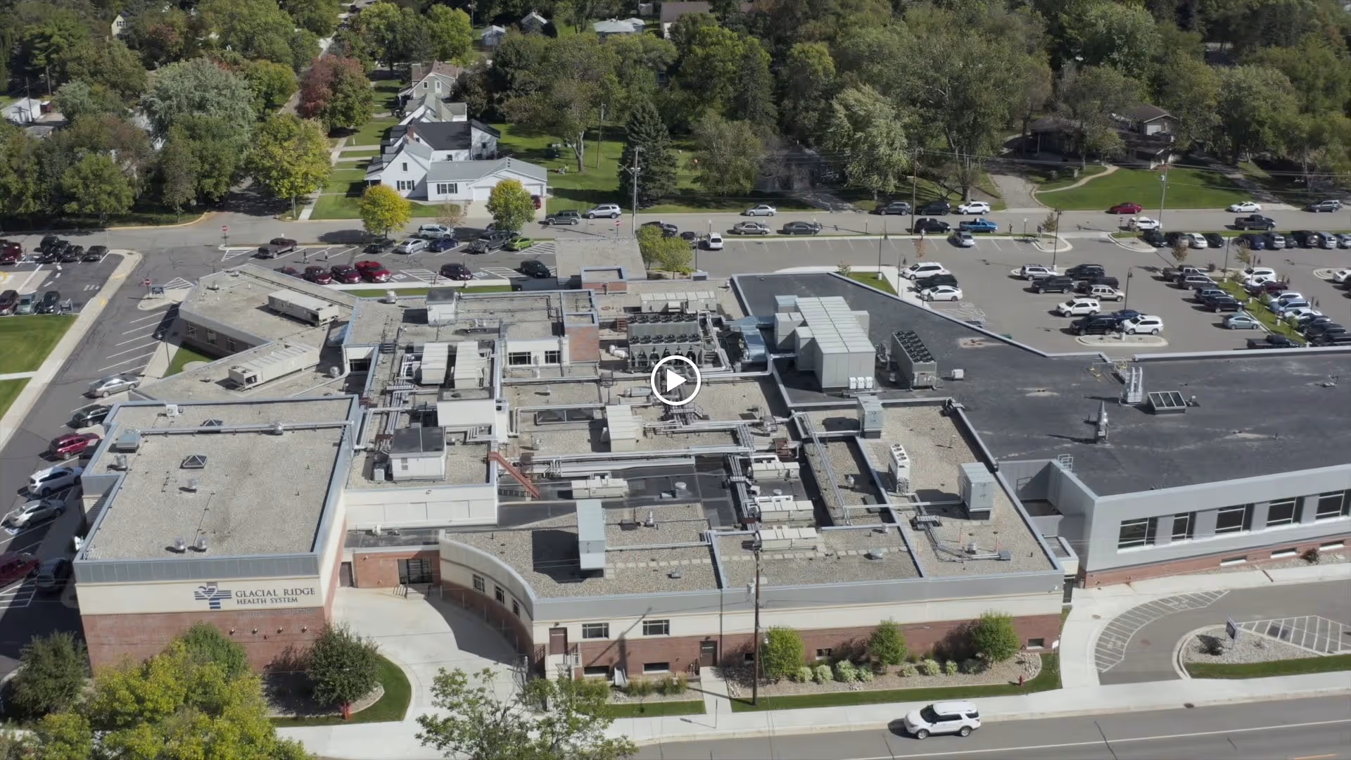 Aerial view of a large health facility with flat roofs and rooftop HVAC units, entrance drive, parking lots, and surrounding trees.