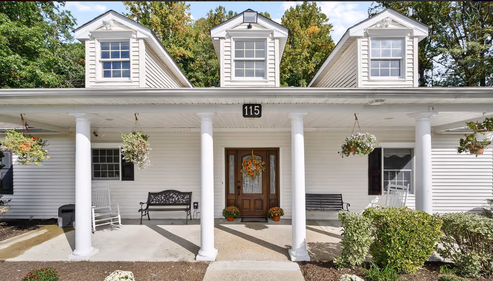 Front exterior view of a white building with three dormer windows on the roof, a covered porch supported by four white columns, hanging flower baskets, benches, rocking chairs, and a wooden door decorated with a wreath and potted plants on either side. The building number 115 is displayed above the door.