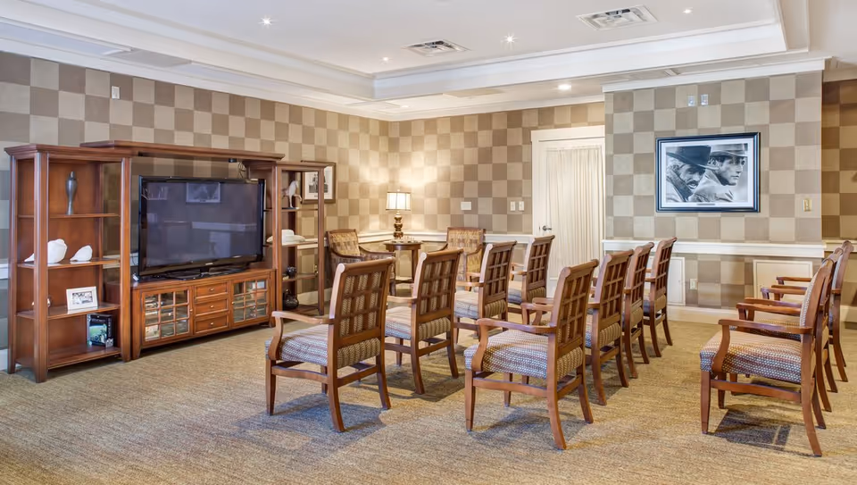 A well-lit room with multiple wooden chairs arranged in rows facing a large flat-screen TV on a wooden entertainment center. The walls have a checkered pattern wallpaper, and there is a framed black and white picture on the wall. A small table with a lamp and two chairs is in the corner.