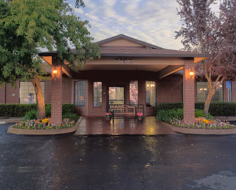 Entrance of Auburn Oaks Care Center featuring a covered porch with brick pillars, a bench, potted flowers, and surrounding greenery including trees and flower beds. The building is made of red brick and has windows on either side of the entrance. The pavement in front is wet and there are lights on the pillars illuminating the area.