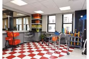 A rehabilitation therapy room with red and white checkered flooring, a red chair at a desk with a computer, exercise equipment including a walker, therapy balls, and colorful weights on racks, with large windows letting in natural light.