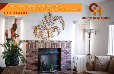 Cozy living room with a brick fireplace decorated with plants and a metal leaf wall art above it. There are two windows with vertical blinds on either side of the fireplace, a tall floor lamp, and comfortable seating with cushions.