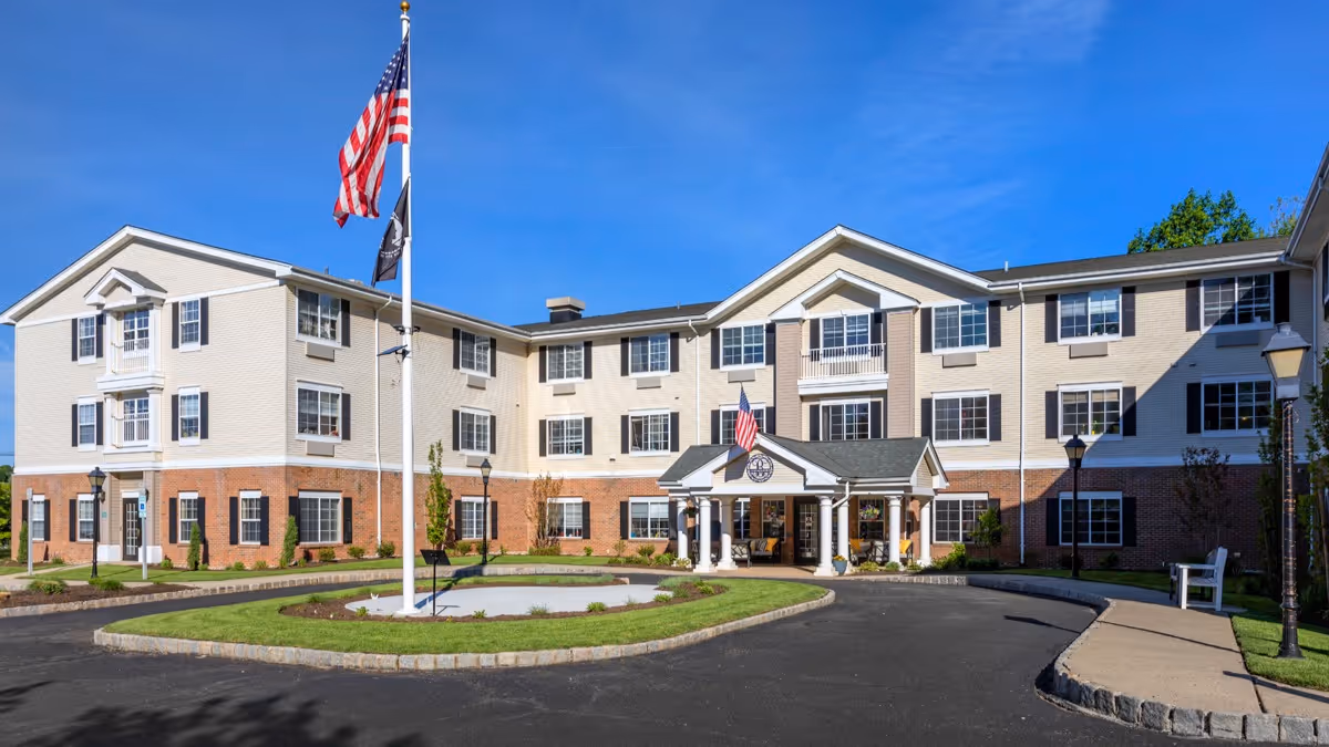 Exterior view of a three-story senior living facility building with beige and brick facade, multiple windows with black shutters, a covered entrance with white columns, and an American flag flying on a flagpole in front. The sky is clear and blue.