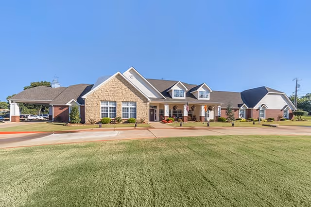 Exterior view of a single-story senior living facility building with a combination of stone and brick facade, multiple windows, a covered entrance with columns, and a well-maintained lawn in front under a clear blue sky.