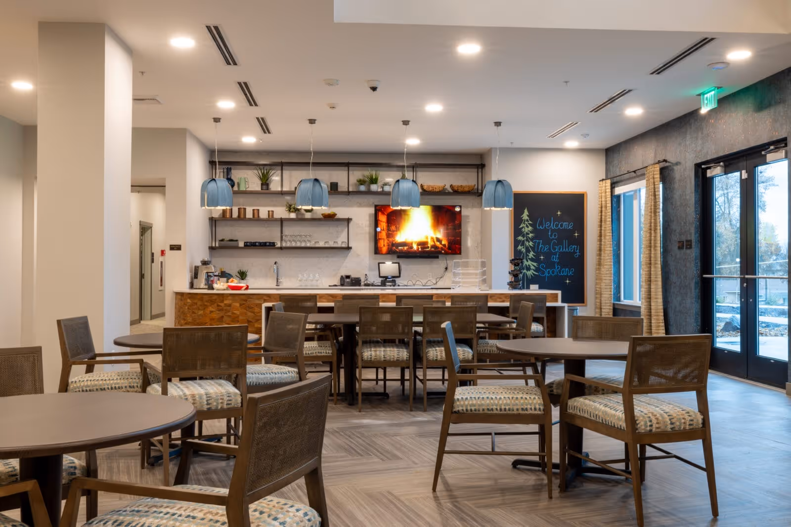 Communal dining and seating area with round tables and chairs, a service counter with pendant lights, a wall-mounted TV showing a fireplace, and a chalkboard welcome sign.