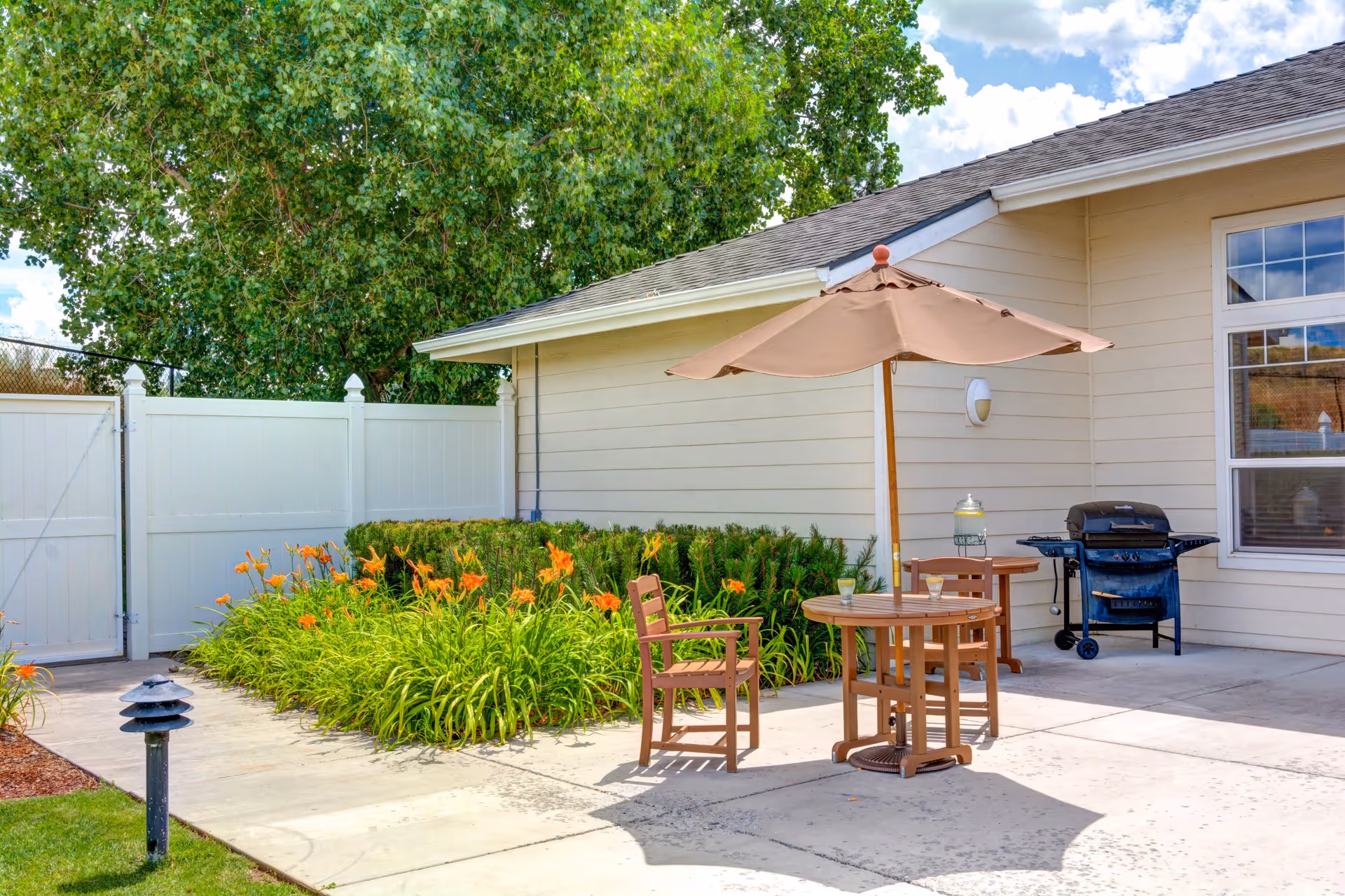 Outdoor patio area with a round wooden table and two chairs under a beige umbrella. There is a barbecue grill near the beige building wall, and a garden with orange flowers and green bushes along the side. A white fence and a large tree are visible in the background under a partly cloudy sky.