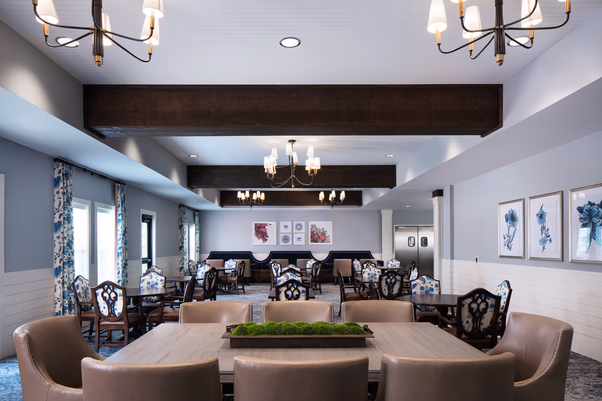 Well-lit communal dining room with multiple tables and chairs, chandeliers, floral artwork, and patterned curtains.