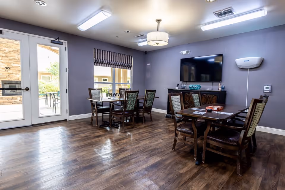 A common area in an assisted living facility with wooden flooring, two tables each surrounded by four chairs, a large flat-screen TV mounted on a purple wall, and a cabinet beneath the TV. There is a window with a striped Roman shade and a glass door leading outside, where patio furniture is visible.