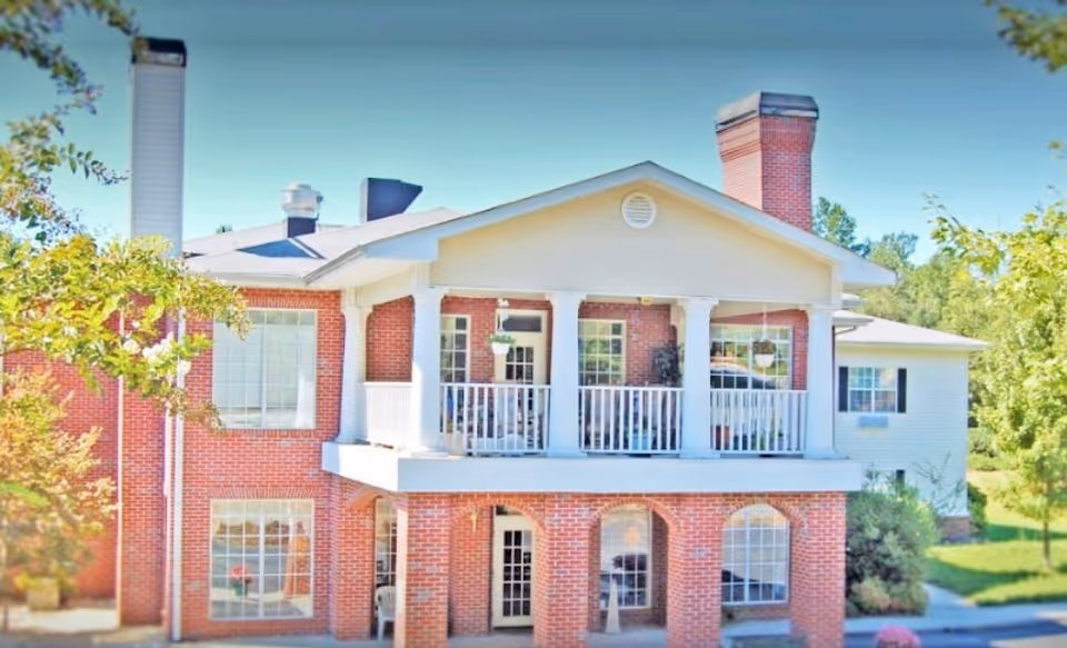 Exterior view of a two-story brick building with white columns and a balcony on the upper floor. The building has large windows, a chimney, and is surrounded by greenery and trees under a clear blue sky.