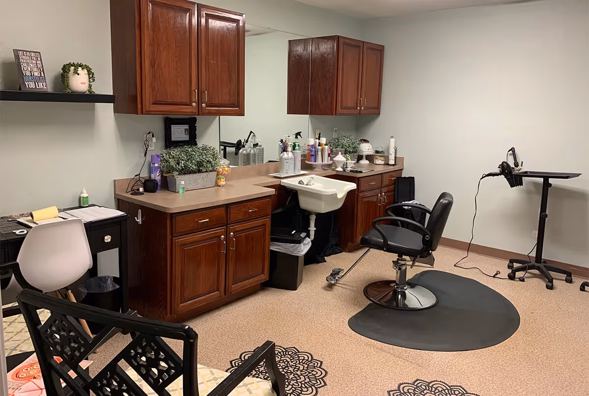 Interior of a hair salon room with a black salon chair on a black mat in front of a white sink. The room has wooden cabinets with various hair care products on the countertop. There is a small desk with a white chair and decorative items including a plant and a framed quote on a shelf above the desk.