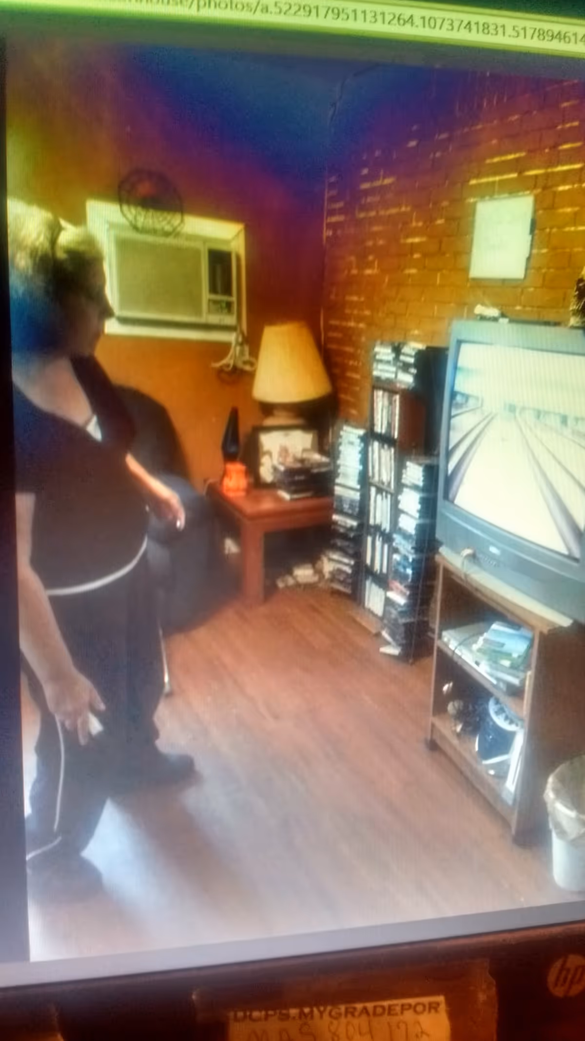 A woman standing in a living room with wooden floors and a brick wall. The room contains a TV on a stand, a small table with a lamp, and shelves filled with DVDs or video games. There is an air conditioning unit mounted on the wall and a dark-colored armchair in the corner.